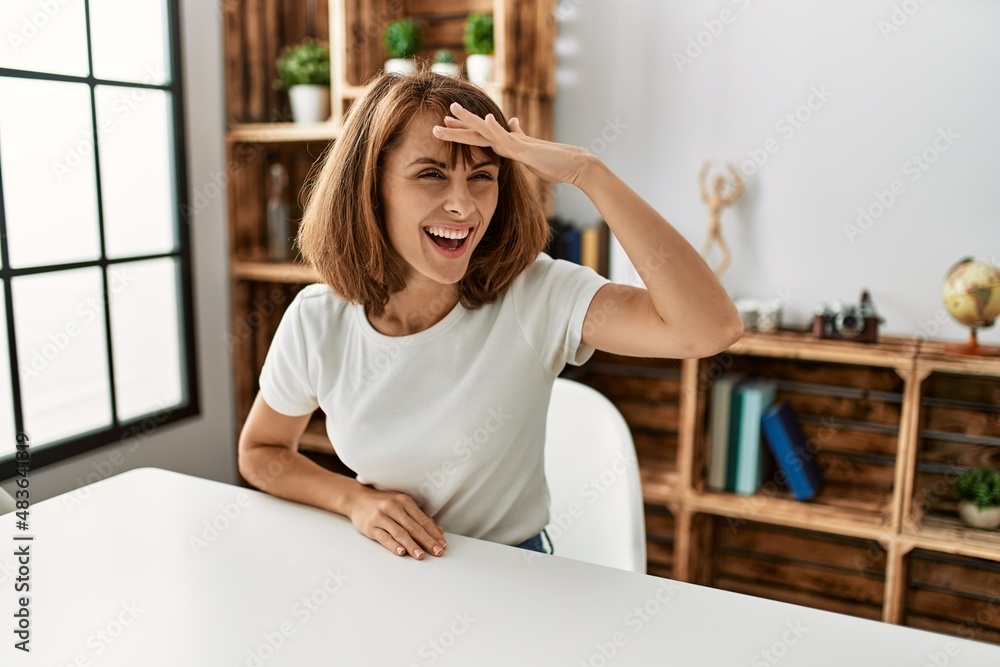 Young caucasian girl wearing casual clothes sitting on the table at home very happy and smiling looking far away with hand over head. searching concept. Young caucasian girl wearing casual clothes sitting on the table at home very happy and smiling looking far away with hand over head. searching concept.