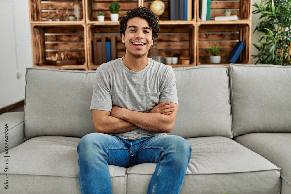 Young hispanic man wearing casual clothes sitting on the sofa at home happy face smiling with crossed arms looking at the camera. positive person. Young hispanic man wearing casual clothes sitting on the sofa at home happy face smiling with crossed arms looking at the camera. positive person.