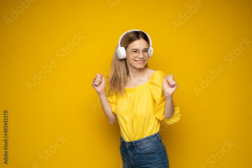 Beautiful young blonde woman with headphones listening to music and smiling and dancing, isolated on yellow background. © Volodymyr