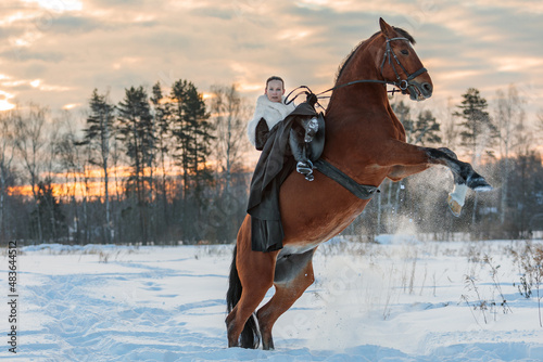 A girl in a white cloak rides a brown horse in winter. © ЮРИЙ ПОЗДНИКОВ