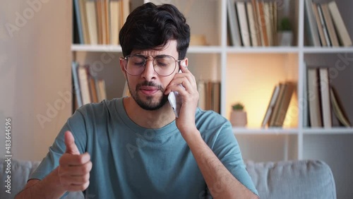 Mobile disappointment. Bad news. Negative communication. Dissatisfied annoyed guy speaking on phone showing refusal thumb down gesture at modern home interior. photo