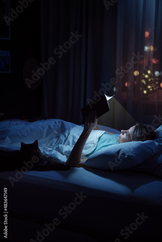 Magic night. Young woman reading book at cozy bed with her cat.