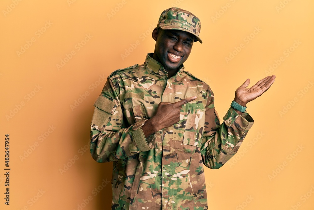 Young african american man wearing army uniform amazed and smiling to the camera while presenting with hand and pointing with finger. Young african american man wearing army uniform amazed and smiling to the camera while presenting with hand and pointing with finger.