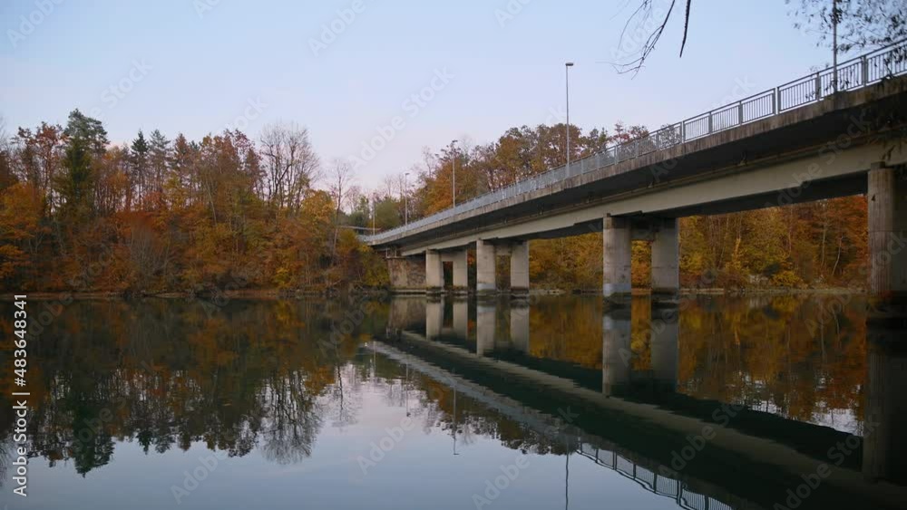 Low angle view of calm Sava River flowing under concrete bridge. Autumn season in Slovenia. Reflection of viaduct in the water. Car traffic on the bridge. Beautiful sunset. Real time, static shot Low angle view of calm Sava River flowing under concrete bridge. Autumn season in Slovenia. Reflection of viaduct in the water. Car traffic on the bridge. Beautiful sunset. Real time, static shot