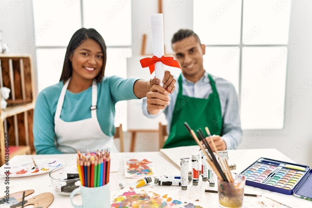 Young latin painter couple smiling happy holding diploma sitting on the table at art studio Young latin painter couple smiling happy holding diploma sitting on the table at art studio