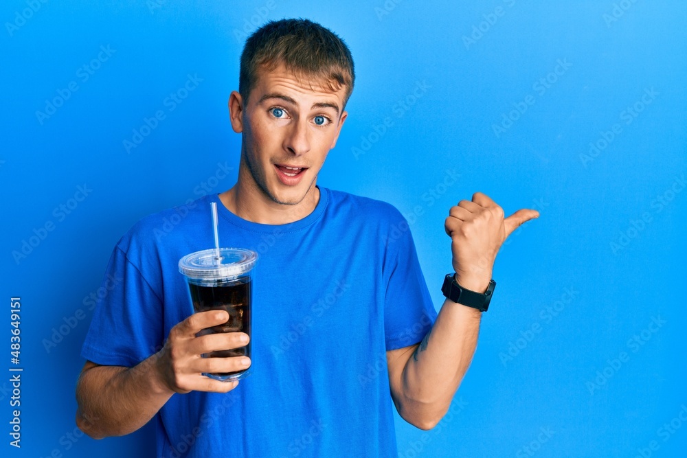 Young caucasian man drinking glass of soda beverage pointing thumb up to the side smiling happy with open mouth Young caucasian man drinking glass of soda beverage pointing thumb up to the side smiling happy with open mouth
