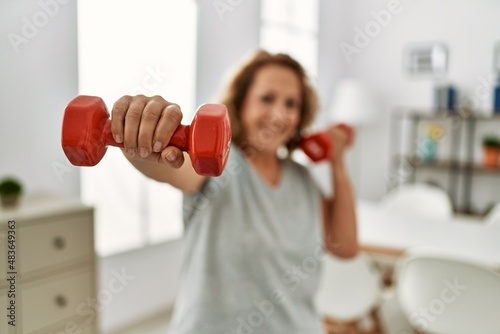 Middle age caucasian woman smiling confident training using dumbbells at home