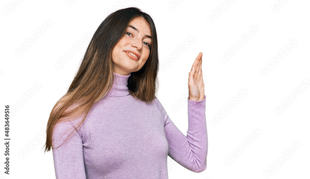Young beautiful teen girl wearing turtleneck sweater waiving saying hello happy and smiling, friendly welcome gesture