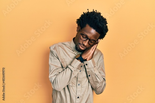 Young african american man wearing casual clothes and glasses sleeping tired dreaming and posing with hands together while smiling with closed eyes.