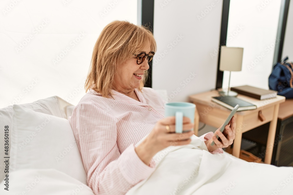 Middle age blonde woman lying on the bed using smartphone and drinking coffee at home. Middle age blonde woman lying on the bed using smartphone and drinking coffee at home.