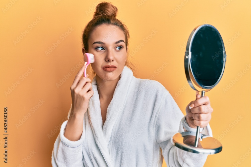 Young blonde woman wearing robe holding makeup remover brush relaxed with serious expression on face. simple and natural looking at the camera. Young blonde woman wearing robe holding makeup remover brush relaxed with serious expression on face. simple and natural looking at the camera.