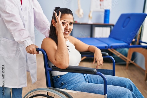 Young hispanic woman sitting on wheelchair at physiotherapy clinic smiling happy doing ok sign with hand on eye looking through fingers