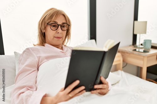 Middle age blonde woman reading book lying on the bed at home.