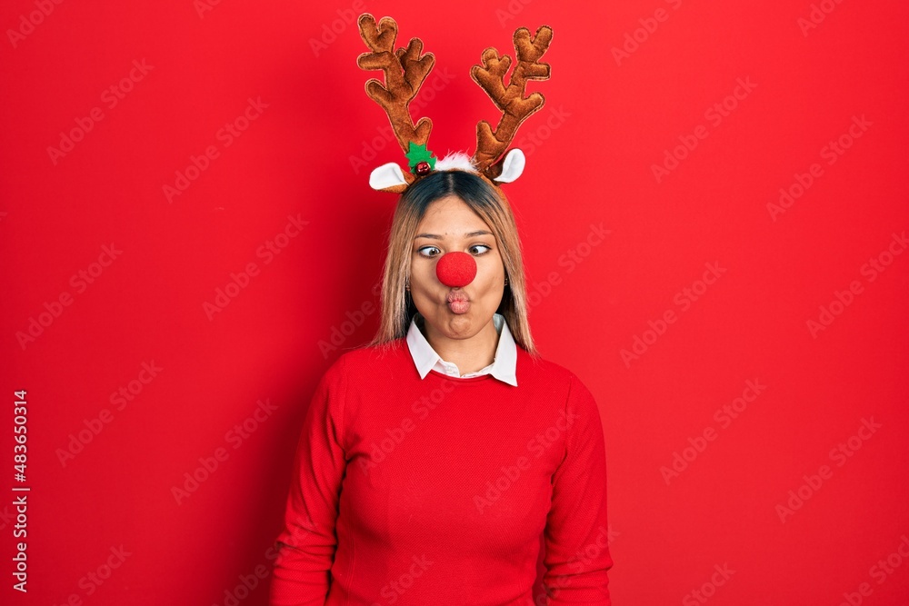 Beautiful hispanic woman wearing deer christmas hat and red nose making fish face with lips, crazy and comical gesture. funny expression. Beautiful hispanic woman wearing deer christmas hat and red nose making fish face with lips, crazy and comical gesture. funny expression.