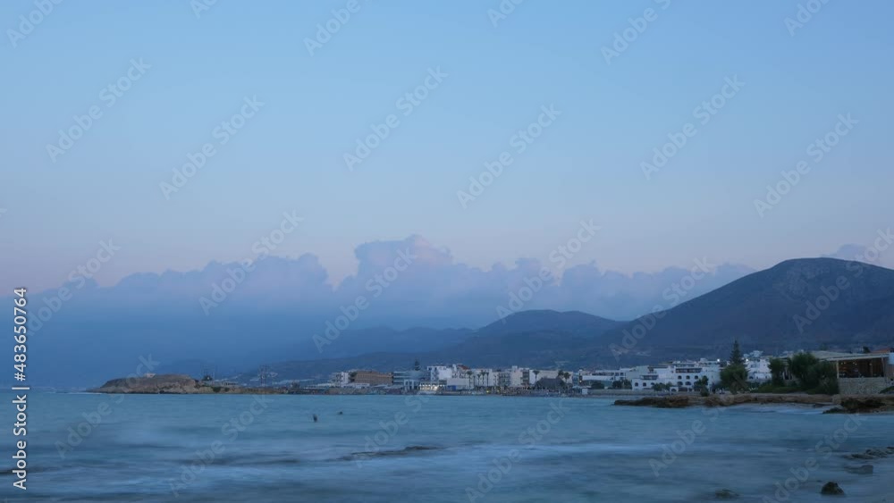 Evening twilight at sea shore, time lapse shot after sunset. Small waves washing beaches of North Crete, small buildings seen on shore. Dim blue hour view Evening twilight at sea shore, time lapse shot after sunset. Small waves washing beaches of North Crete, small buildings seen on shore. Dim blue hour view