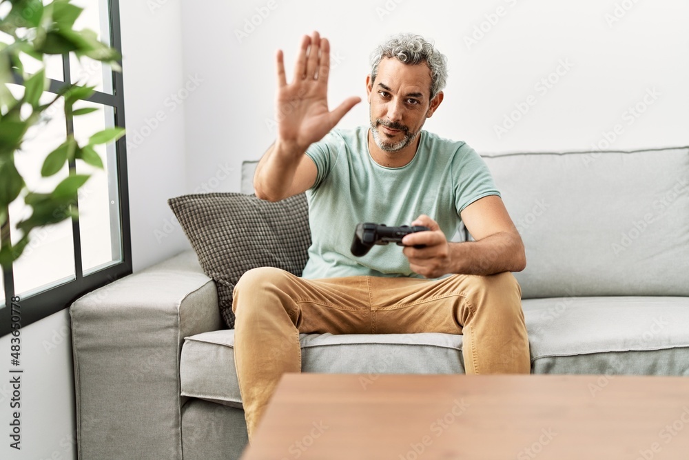 Middle age hispanic man playing video game sitting on the sofa with open hand doing stop sign with serious and confident expression, defense gesture