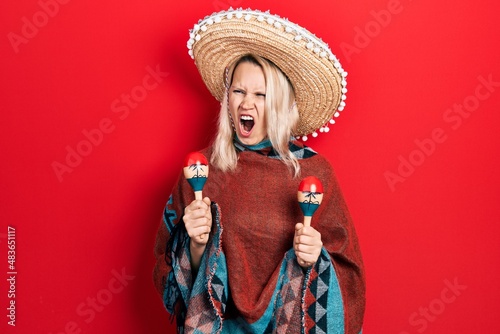 Beautiful caucasian blonde woman wearing festive mexican poncho and maracas angry and mad screaming frustrated and furious, shouting with anger. rage and aggressive concept.