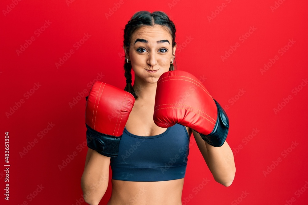 Young brunette girl using boxing gloves puffing cheeks with funny face. mouth inflated with air, catching air.
