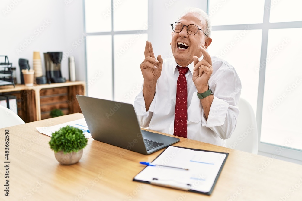 Senior man working at the office using computer laptop gesturing finger crossed smiling with hope and eyes closed. luck and superstitious concept. Senior man working at the office using computer laptop gesturing finger crossed smiling with hope and eyes closed. luck and superstitious concept.