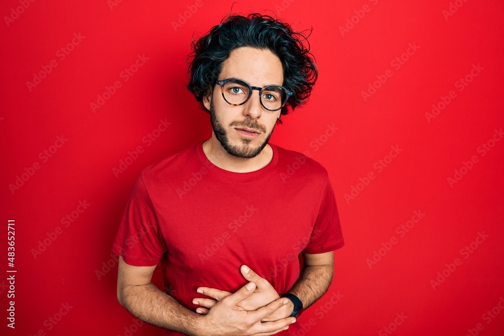 Handsome hispanic man wearing casual t shirt and glasses with hand on stomach because indigestion, painful illness feeling unwell. ache concept.