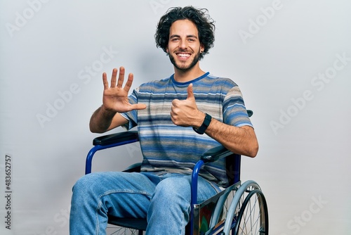 Handsome hispanic man sitting on wheelchair showing and pointing up with fingers number six while smiling confident and happy.