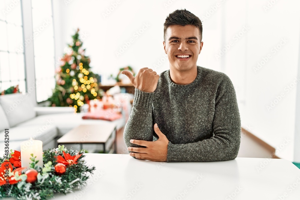 Young hispanic man sitting on the table by christmas tree smiling with happy face looking and pointing to the side with thumb up.