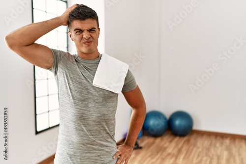 Young hispanic man wearing sportswear and towel at the gym confuse and wonder about question. uncertain with doubt, thinking with hand on head. pensive concept.