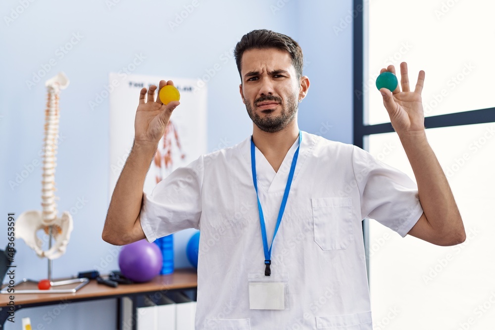 Young hispanic physiotherapist man holding strength balls to train hand muscles clueless and confused expression. doubt concept. Young hispanic physiotherapist man holding strength balls to train hand muscles clueless and confused expression. doubt concept.