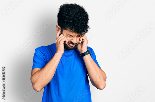 Young arab man with beard wearing casual blue t shirt covering ears with fingers with annoyed expression for the noise of loud music. deaf concept.