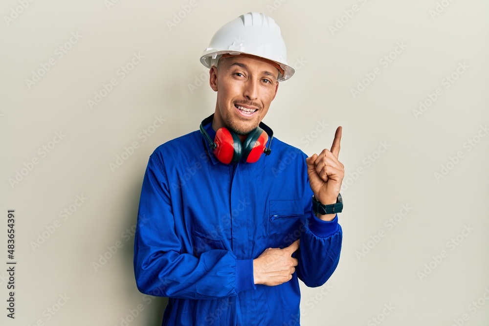 Bald man with beard wearing builder jumpsuit uniform and hardhat smiling happy pointing with hand and finger to the side
