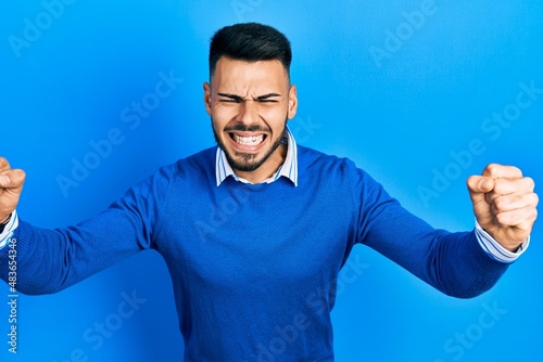 Young hispanic man with beard wearing casual blue sweater angry and mad raising fists frustrated and furious while shouting with anger. rage and aggressive concept.