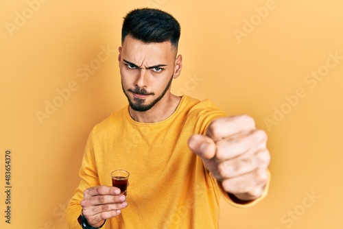 Young hispanic man with beard drinking whiskey shot annoyed and frustrated shouting with anger, yelling crazy with anger and hand raised