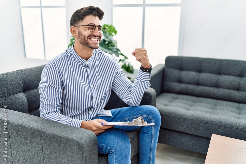 Young psychologist man at consultation office very happy and excited doing winner gesture with arms raised, smiling and screaming for success. celebration concept. Young psychologist man at consultation office very happy and excited doing winner gesture with arms raised, smiling and screaming for success. celebration concept.
