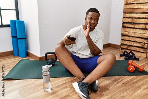 Young african man sitting on training mat at the gym using smartphone hand on mouth telling secret rumor, whispering malicious talk conversation