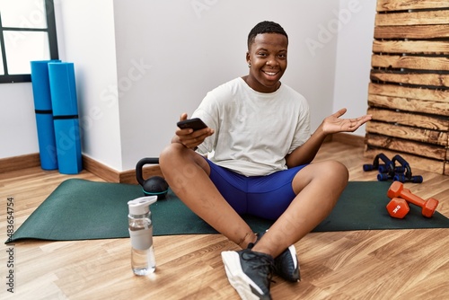 Young african man sitting on training mat at the gym using smartphone pointing aside with hands open palms showing copy space, presenting advertisement smiling excited happy