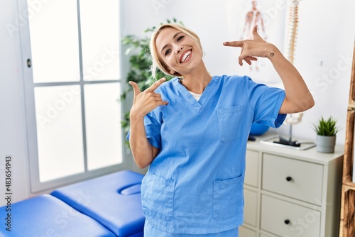 Beautiful blonde physiotherapist woman working at pain recovery clinic smiling cheerful showing and pointing with fingers teeth and mouth. dental health concept.