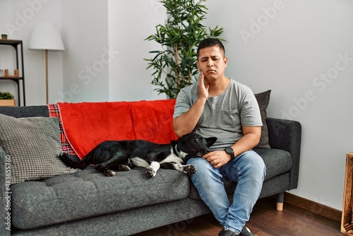 Young latin man and dog sitting on the sofa at home touching mouth with hand with painful expression because of toothache or dental illness on teeth. dentist © Krakenimages.com