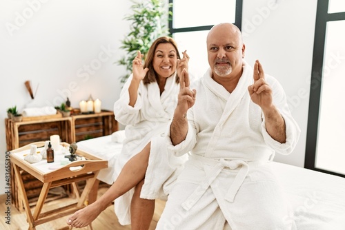 Middle age hispanic couple wearing bathrobe at wellness spa gesturing finger crossed smiling with hope and eyes closed. luck and superstitious concept.