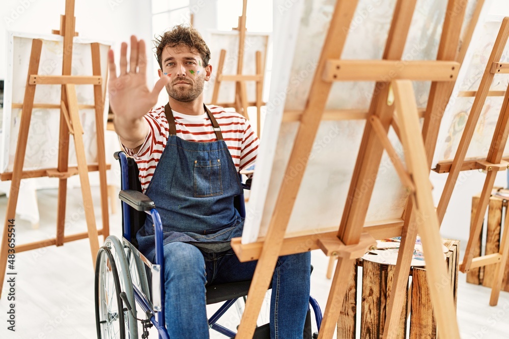 Young handsome man with beard at art studio sitting on wheelchair with open hand doing stop sign with serious and confident expression, defense gesture Young handsome man with beard at art studio sitting on wheelchair with open hand doing stop sign with serious and confident expression, defense gesture