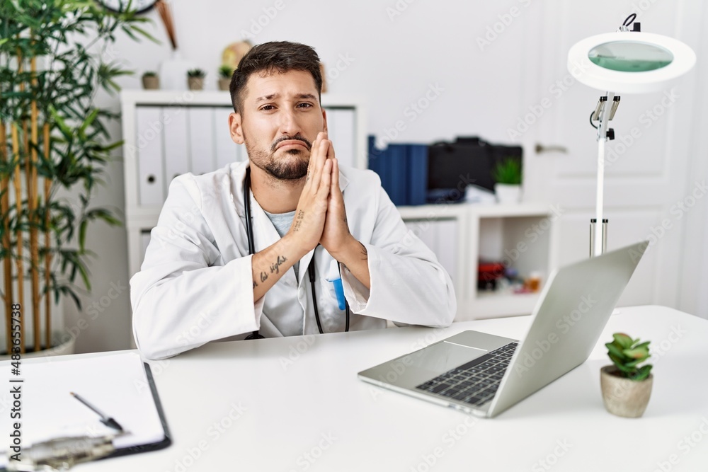 Young doctor working at the clinic using computer laptop begging and praying with hands together with hope expression on face very emotional and worried. begging.