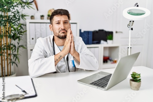 Young doctor working at the clinic using computer laptop begging and praying with hands together with hope expression on face very emotional and worried. begging.