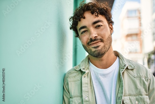 Handsome hispanic man with beard smiling happy outdoors on a sunny day leaning on a wall