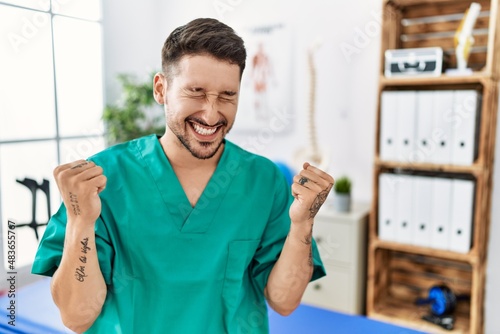 Young physiotherapist man working at pain recovery clinic very happy and excited doing winner gesture with arms raised  smiling and screaming for success. celebration concept.