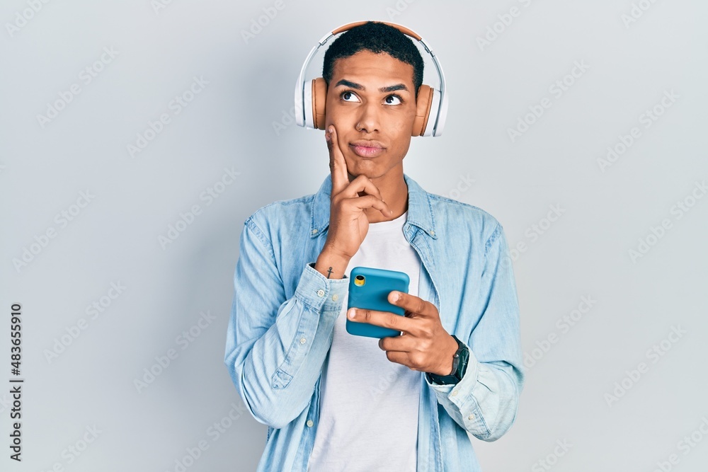 Young african american guy using smartphone wearing headphones serious face thinking about question with hand on chin, thoughtful about confusing idea Young african american guy using smartphone wearing headphones serious face thinking about question with hand on chin, thoughtful about confusing idea