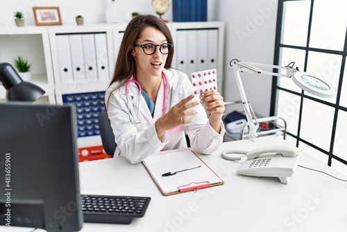 Young hispanic woman wearing doctor uniform holding pills at clinic