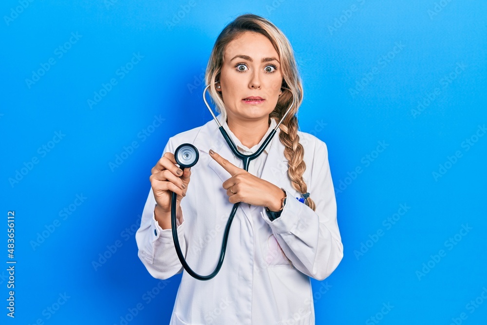 Beautiful young blonde doctor woman holding stethoscope pointing aside worried and nervous with forefinger, concerned and surprised expression Beautiful young blonde doctor woman holding stethoscope pointing aside worried and nervous with forefinger, concerned and surprised expression