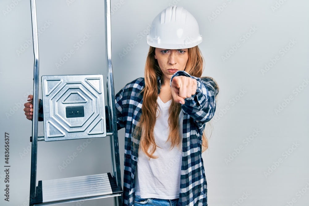 Young caucasian woman wearing hardhat holding ladder pointing with finger to the camera and to you, confident gesture looking serious Young caucasian woman wearing hardhat holding ladder pointing with finger to the camera and to you, confident gesture looking serious
