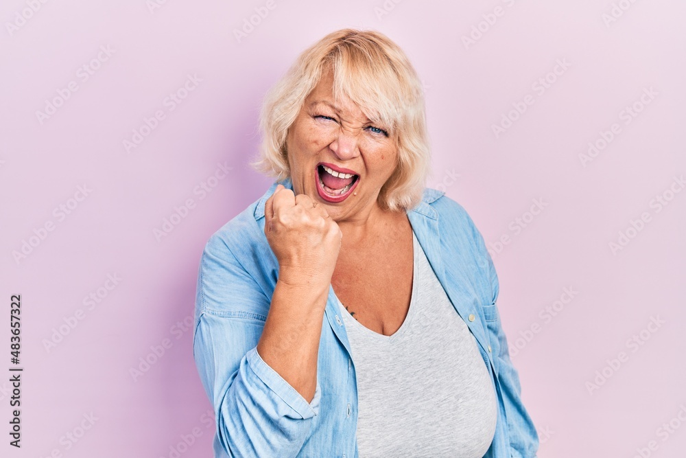 Middle age blonde woman wearing casual clothes angry and mad raising fist frustrated and furious while shouting with anger. rage and aggressive concept. Middle age blonde woman wearing casual clothes angry and mad raising fist frustrated and furious while shouting with anger. rage and aggressive concept.