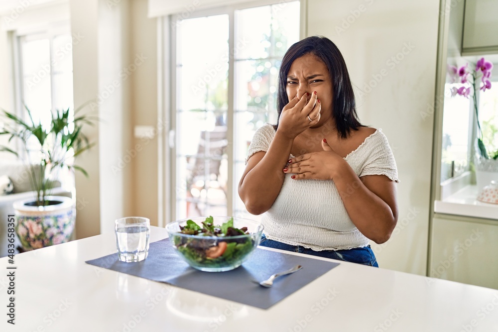 Young hispanic woman eating healthy salad at home smelling something stinky and disgusting, intolerable smell, holding breath with fingers on nose. bad smell