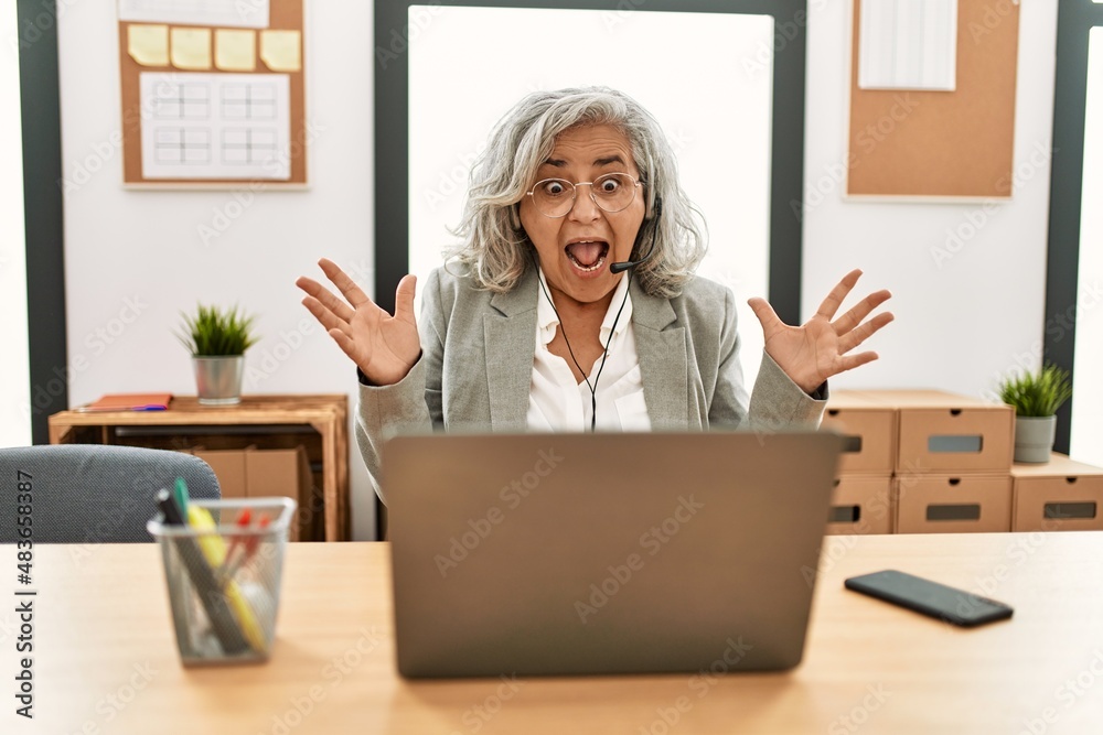 Middle age businesswoman sitting on desk working using laptop at office celebrating crazy and amazed for success with arms raised and open eyes screaming excited. winner concept Middle age businesswoman sitting on desk working using laptop at office celebrating crazy and amazed for success with arms raised and open eyes screaming excited. winner concept